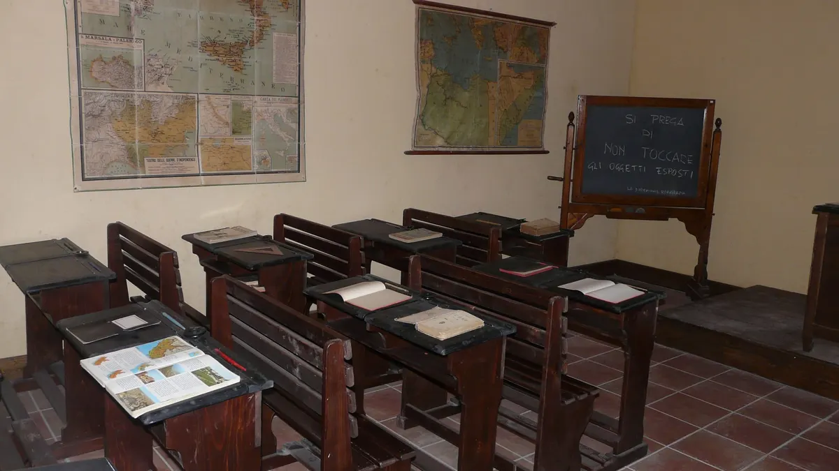 Aula scolastica d'epoca con banchi di legno, carte geografiche alle pareti e una lavagna