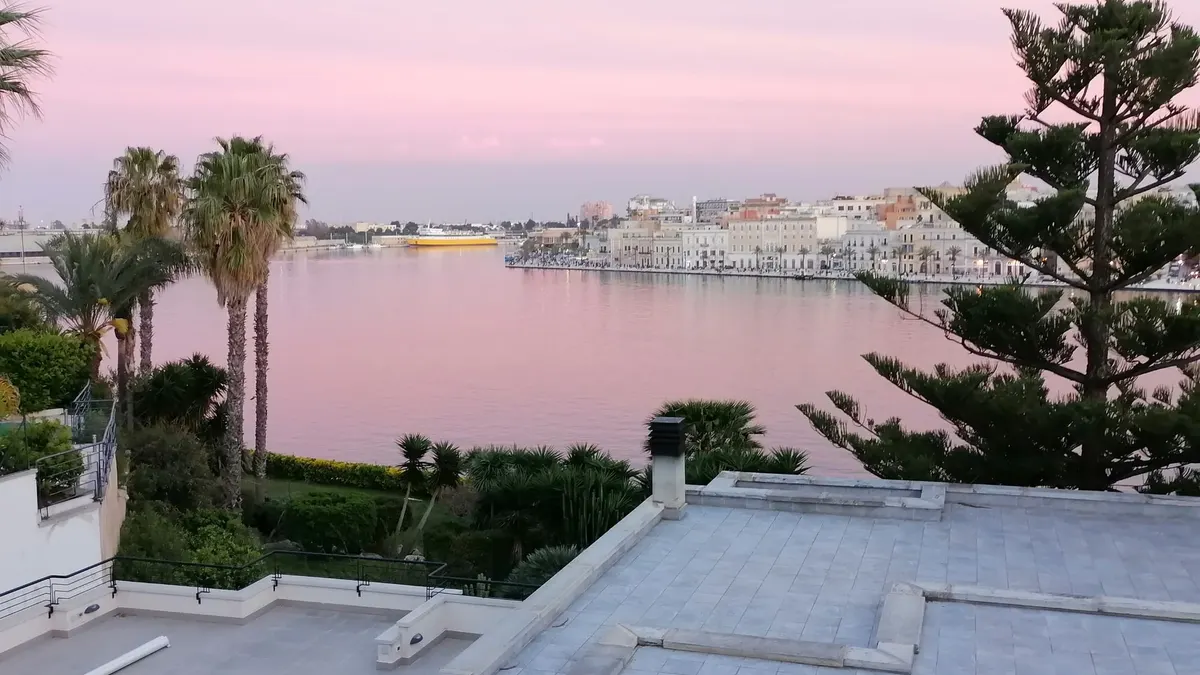 Veduta del porto di Brindisi al tramonto, con l'acqua rosata, palme e le case sul lungomare